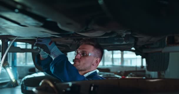 Colpo scorrevole di giovane caucasico bello in uniforme e occhiali in piedi sotto l'auto e torcendo qualche dettaglio con chiave inglese in garage. Meccanico di sesso maschile che lavora in autofficina e riparazioni . — Video Stock