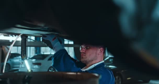 Vista sobre la rueda en el hombre caucásico en uniforme y gafas de pie debajo del coche y detalle de torsión con llave inglesa en el garaje. Mecánico masculino que trabaja duro en el servicio automático y el sistema de roturas de reparación. Disparo deslizante . — Vídeo de stock