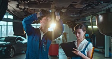 Beyaz bir adam, genç bir kadına araba arızalarını tamir etmeyi öğretiyor ve otomobil servisinde teknik inceleme gösteriyor. Kadın işçi öğrenim ve not alırken, erkek meslektaşı da yardım ve gösteri yapıyor..