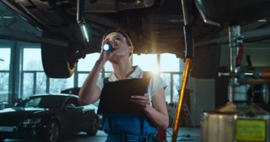 Beyaz kadın arabanın altında duruyor ve garajda el feneriyle kontrol ediyor. Otomobil servisinde çalışan ve otomobilin teknik incelemesini yapan kadın tamirci. Belge dolduran ve not alan işçiler.