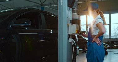 Üniformalı ve şapkalı beyaz bir kadın oto hizmet ve onarım işinde çalışıyor. Kadın işçi tamire başlamak için düğmeye basıyor ve arabayı kaldırıyor..