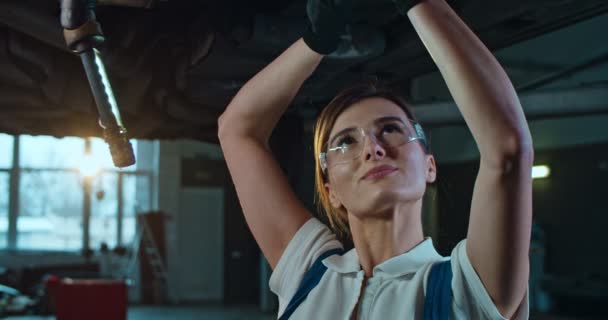Närbild av vit ung vacker kvinna i uniform och glasögon står under bilen och vrider några detaljer med skiftnyckel i garage. Kvinnlig mekaniker arbetar i auto service salong och reparationsraster. — Stockvideo