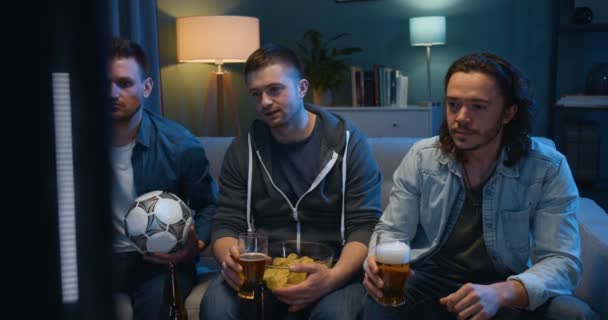 Drie Kaukasische jongens zitten op de bank in de donkere woonkamer met voetbal en chips en kijken naar voetbalwedstrijd op tv Beste mannelijke vrienden besteden avond met sportkanaal en doen proost met bier — Stockvideo