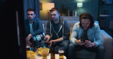 Akşamları kanepede dinlenen ve abur cubur yiyip telefonda oynarken spor televizyon izleyen üç beyaz adam. Evdeki kanepede televizyon karşısında oturan adamlar..