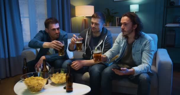 Tre kaukasiska killar tar sina glas med öl och jublar när de sitter på soffan sent på kvällen hemma. Unga män tittar på TV tillsammans på natten med snacks. — Stockvideo