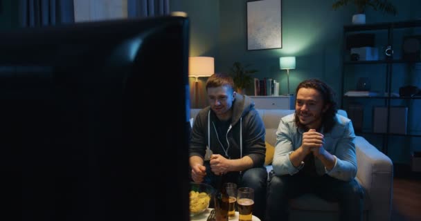 Schöne kaukasische Männerfreunde sitzen auf dem Sofa und sorgen sich um die Fußballmannschaft im Spiel, während sie Sportkanäle im Fernsehen schauen. Torraumkonzept. — Stockvideo