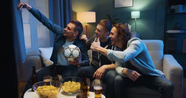 Τρεις όμορφοι λευκοί κολλητοί κάθονται στον καναπέ το βράδυ με μπάλα ποδοσφαίρου, σνακ και μπύρα και βγάζουν selfie φωτογραφία με κάμερα smartphone. Οι φίλαθλοι ποζάρουν στο τηλέφωνο στο σπίτι στην τηλεόραση. — Αρχείο Βίντεο