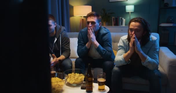 Három kaukázusi, szomorú és aggódó srác, akik otthon ülnek a kanapén, sötét nappaliban, és focimeccset néznek. Kedvenc csapat vesztes koncepció. Felkent férfiak a sportcsatorna előtt.. — Stock videók
