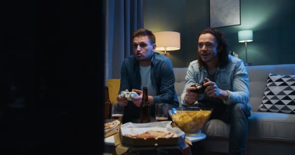 Όμορφοι Καυκάσιοι χαρούμενοι άνδρες φίλοι κάθονται στον καναπέ σε ένταση και ανησυχούν ενώ παίζουν βιντεοπαιχνίδι με joysticks στην οθόνη της τηλεόρασης. Videogaming στο σπίτι έννοια. — Αρχείο Βίντεο