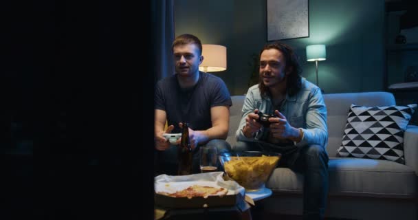 Twee blanke beste mannelijke vrienden die videogame spelen met joystick terwijl ze 's avonds laat op de bank zitten in de woonkamer met snacks en bier voor de tv. Mannen hebben plezier samen met video games. — Stockvideo