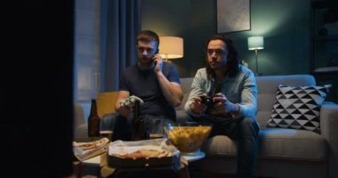 İki Kafkasyalı arkadaş, karanlık bir salonda oturmuş cep telefonuyla konuşan TV ekranı önünde abur cubur ve bira içerken joystick ile video oyunu oynuyor. Erkekler oyunlarla eğleniyor.