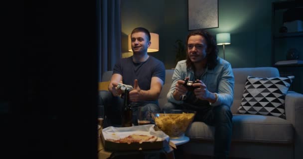 Twee Kaukasische vriendelijke jongens die videogame spelen met joystick terwijl ze 's avonds laat in spanning zitten met snacks en bier voor de tv. Mannen hebben plezier samen met games. — Stockvideo