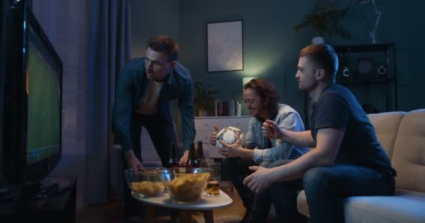 Muž přináší pizzu v tmavém obýváku, kde jeho nejlepší přátelé sedí na pohovce a dívají se na fotbal v televizi s pivem. Kluci jásají pro oblíbený tým pozdě v noci doma. — Stock video
