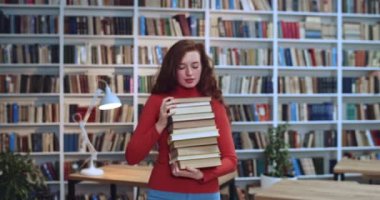 Uzun kıvırcık saçlı, çilli, elinde kitap sayan ve kütüphanede kameraya gülümseyen inek kızıl saçlı kız öğrencinin yakın plan portresi. Arkaplanda kitaplık.