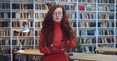 Kollarını kavuşturup kızgın bir ifadeyle kameraya bakan gözlüklü genç kızıl saçlı kızın portresi. Arkaplanda kitaplık.