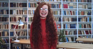 Uzun kıvırcık doğal saçlı ve gözlüklü çekici kızıl saçlı öğrencinin portresi kütüphanede duruyor ve gülümsüyor. Arkaplanda kitaplık.