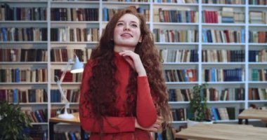 Uzun kıvırcık saçlı, uzun kıvırcık saçlı, kütüphanede flört eden ve kameraya bakan kızıl saçlı bir kadının portresi. Arkaplanda kitaplık.