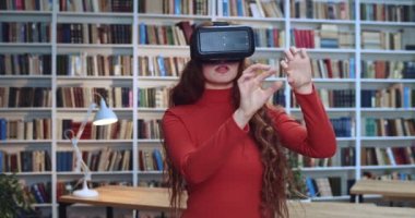 Sanal gerçeklik kulaklığı takan güzel kızıl saçlı kadının yakın plan portresi kütüphanedeki kitaplığın yanındaki modern 3D VR gözlükleri kullanarak bir şeye dokunuyor..