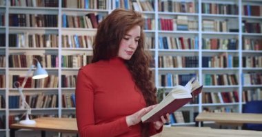Kızıl saçlı inek kızın yakın plan portresi son test için hazırlanıyor ve kütüphanede kitap okuyor. Arkaplanda kitaplık.
