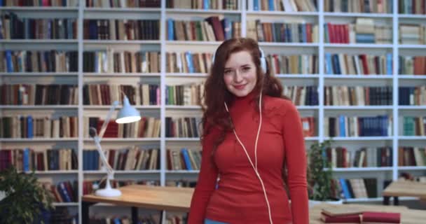 Retrato de dulce hermosa estudiante de cabeza roja con increíbles pecas de pelo largo rizado natural y auriculares blancos bailando y divirtiéndose en la biblioteca. Librería en segundo plano . — Vídeo de stock