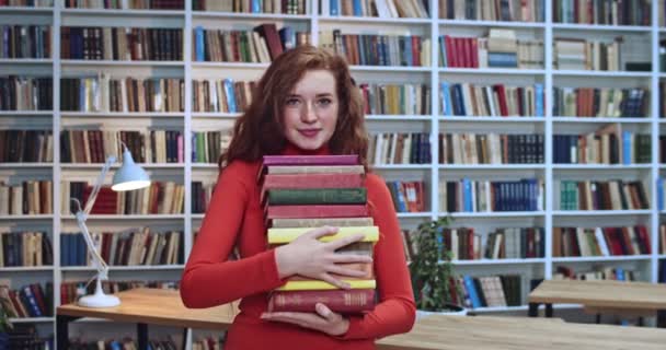 Retrato de menina cabeça vermelha bonito com cabelos longos e cacheados naturais e sardas carregando muitos livros na biblioteca. Livraria no fundo . — Vídeo de Stock