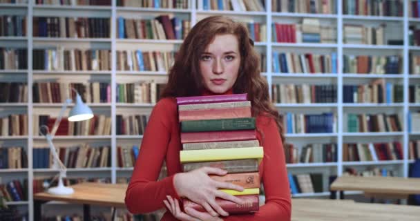 Zbliżenie portret poważnej rudej uczennicy z długimi kręconymi naturalnymi włosami i piegami patrzącymi w kamerę, zachowując wiele książek w bibliotece. Regał na książki w tle. — Wideo stockowe
