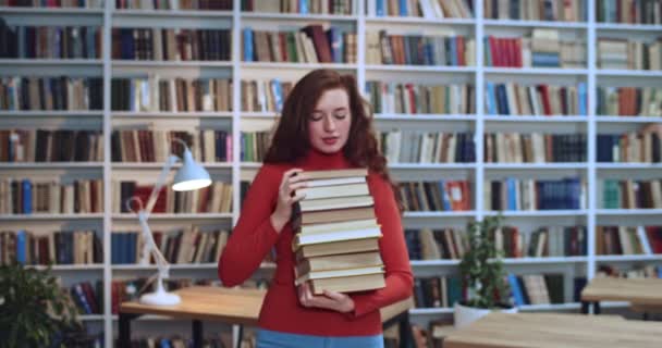 Portrait en gros plan d'une étudiante rousse à la tête rouge avec de longs cheveux naturels bouclés et des taches de rousseur comptant des livres dans ses mains et souriant à la caméra à la bibliothèque. Bibliothèque en arrière-plan . — Video