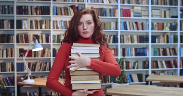 Портрет грустной рыжей женщины с длинными вьющимися натуральными волосами и веснушками, безрадостно смотрящей в камеру и несущей книги в библиотеке. Книжный шкаф в фоновом режиме . — стоковое видео