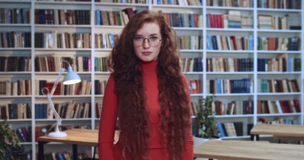 Portret van een getalenteerde jonge roodharige studente met een bril die haar armen kruist en met een serieuze gezichtsuitdrukking naar de camera kijkt. Boekenkast op de achtergrond. — Stockvideo