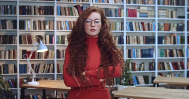 Retrato de jovem talentosa cabeça vermelha menina em óculos cruzando os braços e olhando para a câmera com expressão facial irritada. Livraria no fundo . — Vídeo de Stock