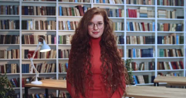 Retrato de una linda estudiante pelirroja con cabello largo y rizado natural y pecas tocando sus gafas y sonriendo en la biblioteca. Librería en segundo plano . — Vídeo de stock