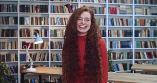 Portret van mooie rode hoofd mooie vrouw met lange krullend natuurlijk haar staan in de bibliotheek en vrolijk spelen met haar haar. Boekenkast op de achtergrond. — Stockvideo