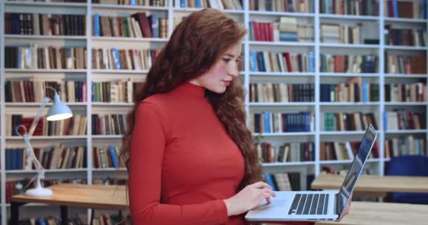 Πορτρέτο της σοβαρής σέξι κόκκινο κεφάλι νεαρή γυναίκα με μακριά σγουρά φυσικά μαλλιά που εργάζονται στη βιβλιοθήκη με υπολογιστή και κοιτάζοντας την οθόνη. Βιβλιοθήκη στο φόντο. — Αρχείο Βίντεο