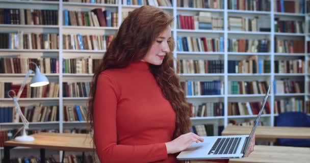 Portret atrakcyjnej rudej bibliotekarki z długimi kręconymi naturalnymi włosami, szukającej w internecie w bibliotece. Regał na książki w tle. — Wideo stockowe