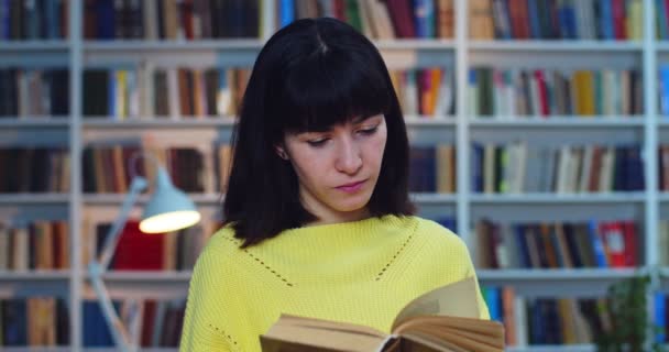 Close-up portret van een lachend brunette meisje met beugels en gele trui ewading boek en staan dicht bij de boekenplank in de bibliotheek — Stockvideo