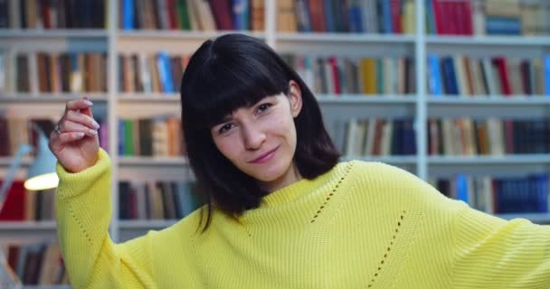 Retrato de cerca de un estudiante atrevido con tirantes en suéter amarillo bailando y divirtiéndose delante de una estantería en la biblioteca — Vídeo de stock