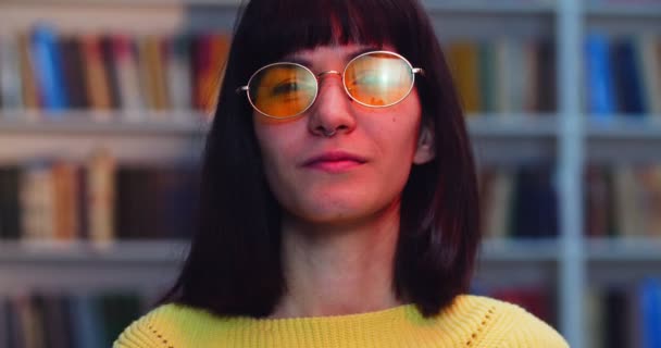 Close-up retrato de uma estudante sorridente em óculos e camisola amarela dançando perto de estante na biblioteca com luzes coloridas . — Vídeo de Stock