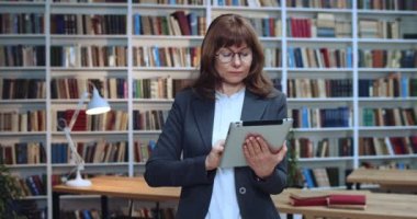 Kütüphanede tablet kullanarak ve e-kitap okuyarak gözlüklü, akıllı Biznesskadının yakın plan portresi. Arkaplanda kitaplık.