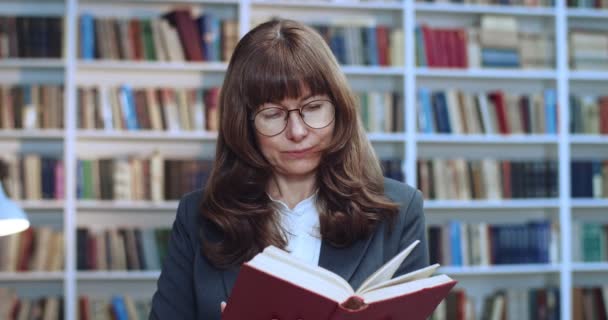 Close-up portrait of brunette female professor in glasses working and reading book in library. Bookcase in background. — Stock Video