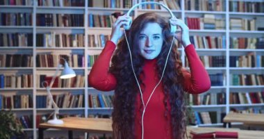 Kızıl saçlı, beyaz kulaklıklarla müzik dinleyen, kütüphanedeki kitaplığın yanında dans eden güzel disko ışıklarıyla dans eden çekici bir kadının yakın plan portresi..