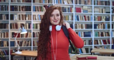 Uzun kıvırcık doğal çilli, beyaz kulaklıklı, sırt çantası takan ve kütüphanede gülümseyen güzel kızıl saçlı kızın portresi. Arkaplanda kitaplık.