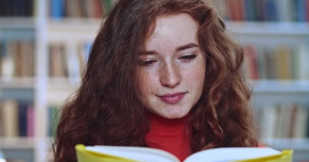 Портрет красивой рыжей головы красивой женщины с длинными вьющимися натуральными волосами, читающей желтые книги и работающей в библиотеке. Книжный шкаф в фоновом режиме . — стоковое видео