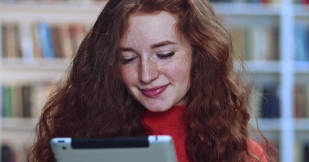 Närbild porträtt av söt röd huvud student med lång lockigt naturligt hår och fräknar med hjälp av surfplatta i biblioteket och läsa e-böcker. Bokhylla i bakgrunden. — Stockvideo