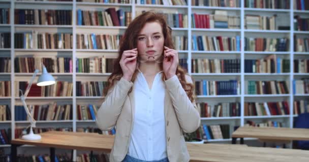 才能ある若い赤頭の女子学生が眼鏡をかけ、真剣な表情でカメラを見ている姿が描かれています。バックグラウンドの本棚. — ストック動画