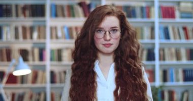 Gözlüklü, gülümseyen ve kameraya bakan güzel kızıl saçlı kıvırcık saçlı kızın portresi. Arkaplanda kitaplık.