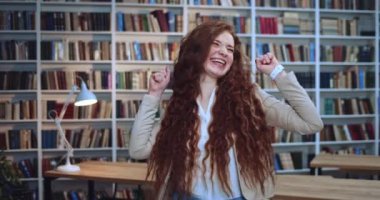 Sınavdan en iyi notu aldıktan sonra kütüphanede dans eden neşeli kızıl saçlı mutlu kız öğrenci Portret. Arkaplanda kitaplık.
