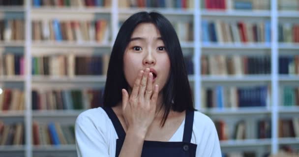 Portret de sorprendida asiática joven estudiante con el pelo negro mirando a la cámara y se cubre la boca con su palma mientras está muy sorprendida en la biblioteca al lado del estante del libro . — Vídeo de stock