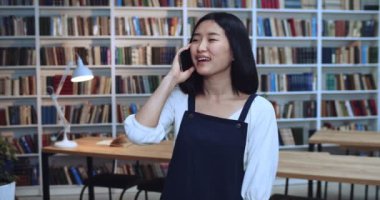 Arkadaş canlısı Asyalı güzel doğal saçlı bir öğrencinin portresi kütüphanede siyah telefonunu kullanan bir arkadaşıyla konuşuyor. Arkaplanda kitaplık.