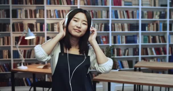 Portret słodkiej pięknej brunetki azjatyckiej studentki z niesamowitymi naturalnymi włosami i białymi słuchawkami tańczącymi i cieszącymi się jej czasem w bibliotece w neonowych światłach. Regał na książki w tle. — Wideo stockowe