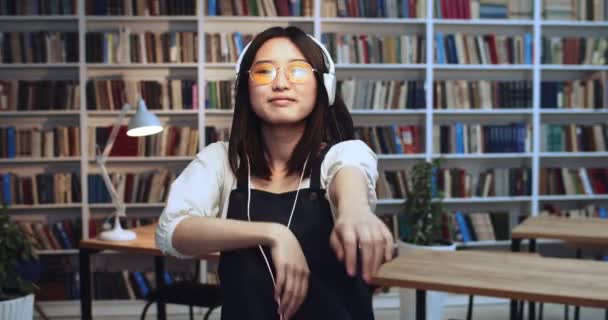 Parlak esmer Asyalı öğrencinin portresi. İnanılmaz doğal saçları ve beyaz kulaklıkları var. Renkli neon ışıklarla kütüphanede dans ediyor. Arkaplanda kitaplık. — Stok video